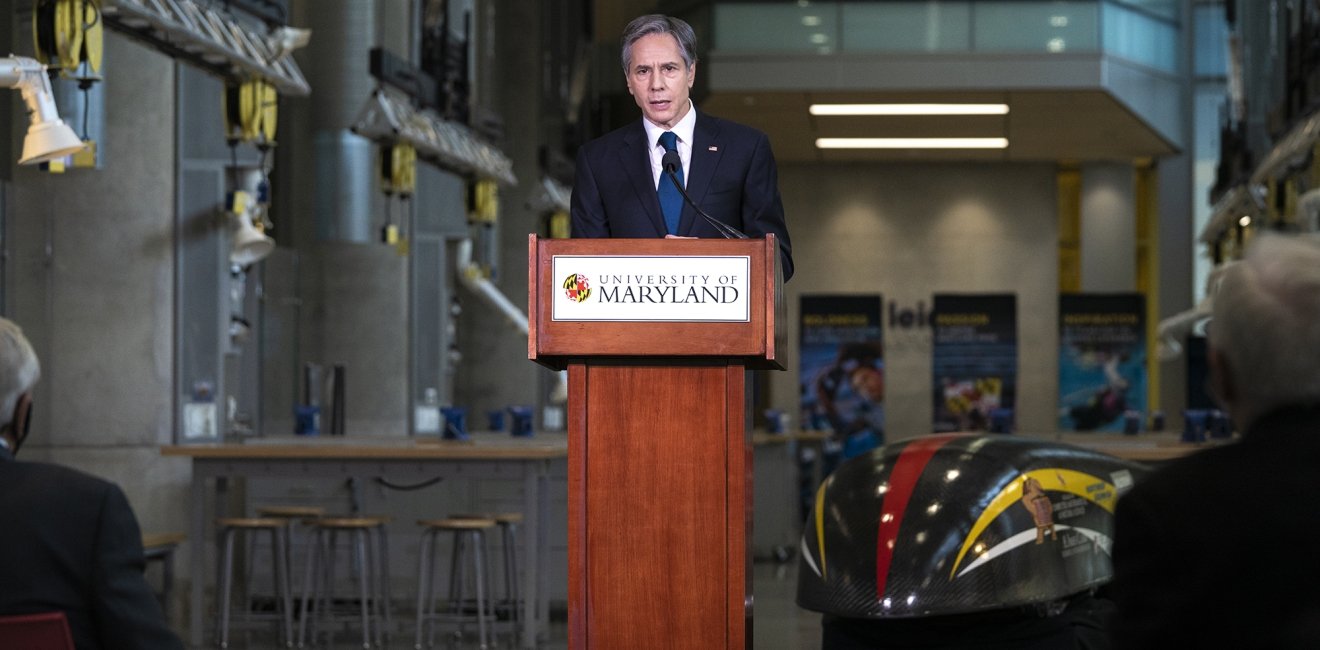 Secretary of State Antony Blinken stands at a podium with a University of Maryland logo on the front, delivering a speech.