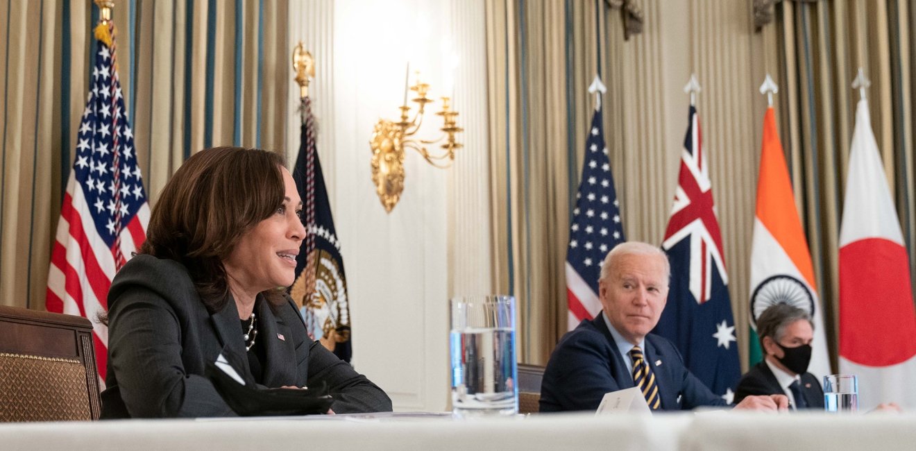 Harris, Biden, and Blinken sit at a table in front of the flags of the United States, Australia, India, and Japan.