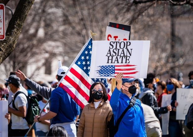 A man stands in a group of protestors wearing a masks, holding a sign with an American flag on it that says Stop Asian Hate.