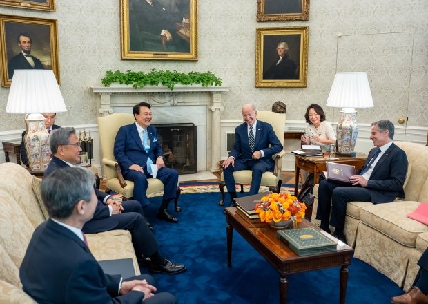 A group of diplomats surrounding Presidents Yoon and Biden in the Oval Office, the group is laughing.