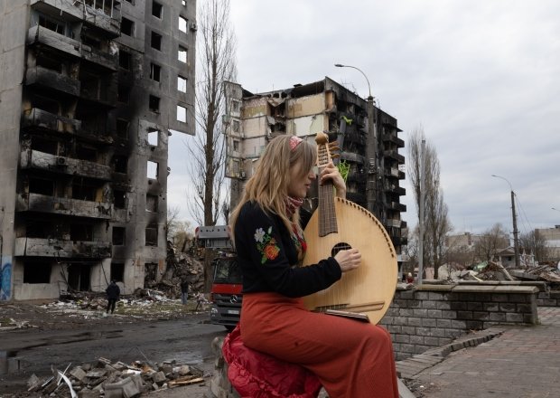 A woman plays a stringed instrument in front of a destroyed building in Bodoryanca, Kyiv Oblast, Ukraine