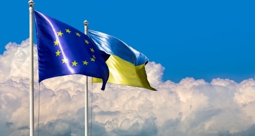 Two flags of Ukraine and the European Union on flagpoles at blue cloudy sky.