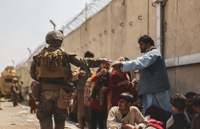 A man in a military uniform hands a bottle of water to an Afghan civilian waiting in a crowd of people.