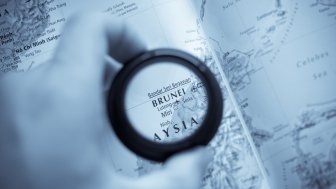 A hand holds a magnifying glass that is circling Brunei on a map.
