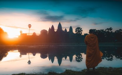 A monk stands in the foreground looking out at a temple at sunrise.
