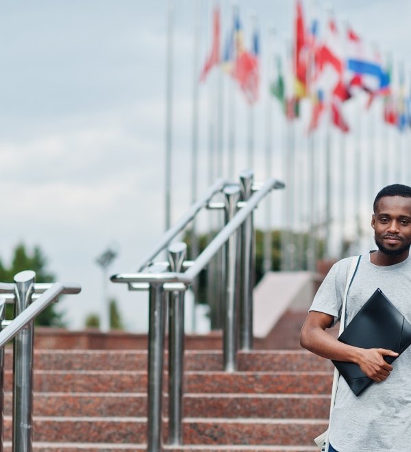 Black student posed with backpack and school items on yard of university, against flags of different countries.