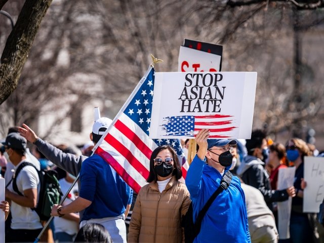 A man stands in a group of protestors wearing a masks, holding a sign with an American flag on it that says Stop Asian Hate.