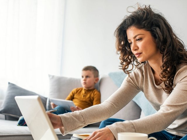 Smiling mom working at home with her child on the sofa while writing an email. 