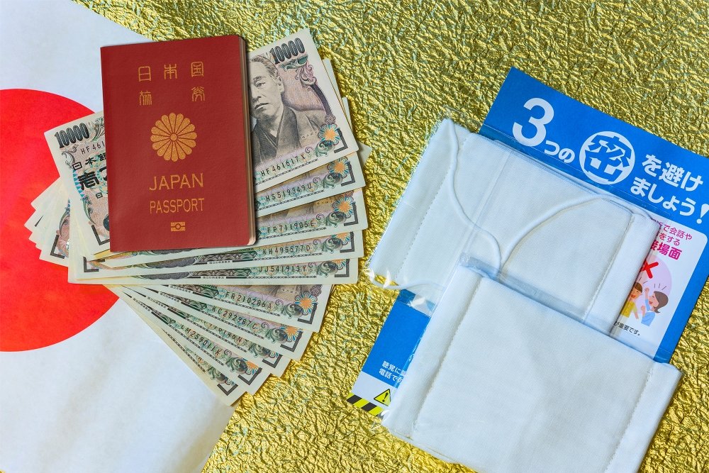 Cloth masks, a leaflet, and 100,000 yen cash sitting on a background with a Japanese flag.