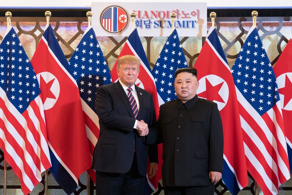 Donald J. Trump stands next to Kim Jong Un as they shake hands and pose in front of U.S. and North Korean flags