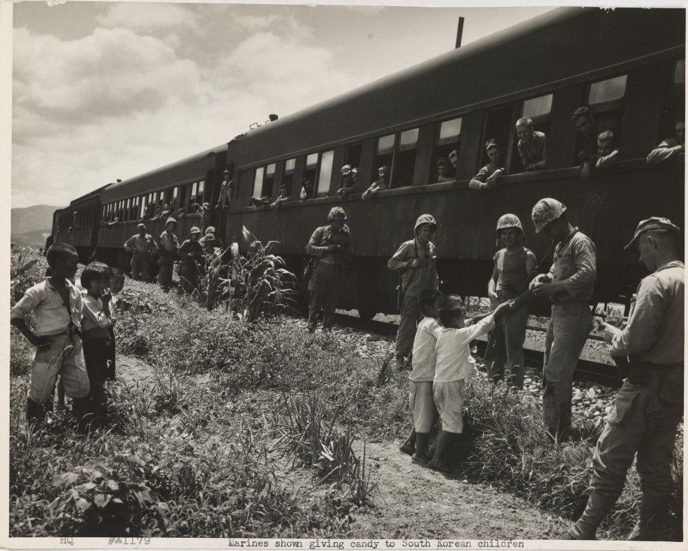 A black and white photo of U.S. Soldiers standing next to a stopped train, handing out candy to South Korean children.