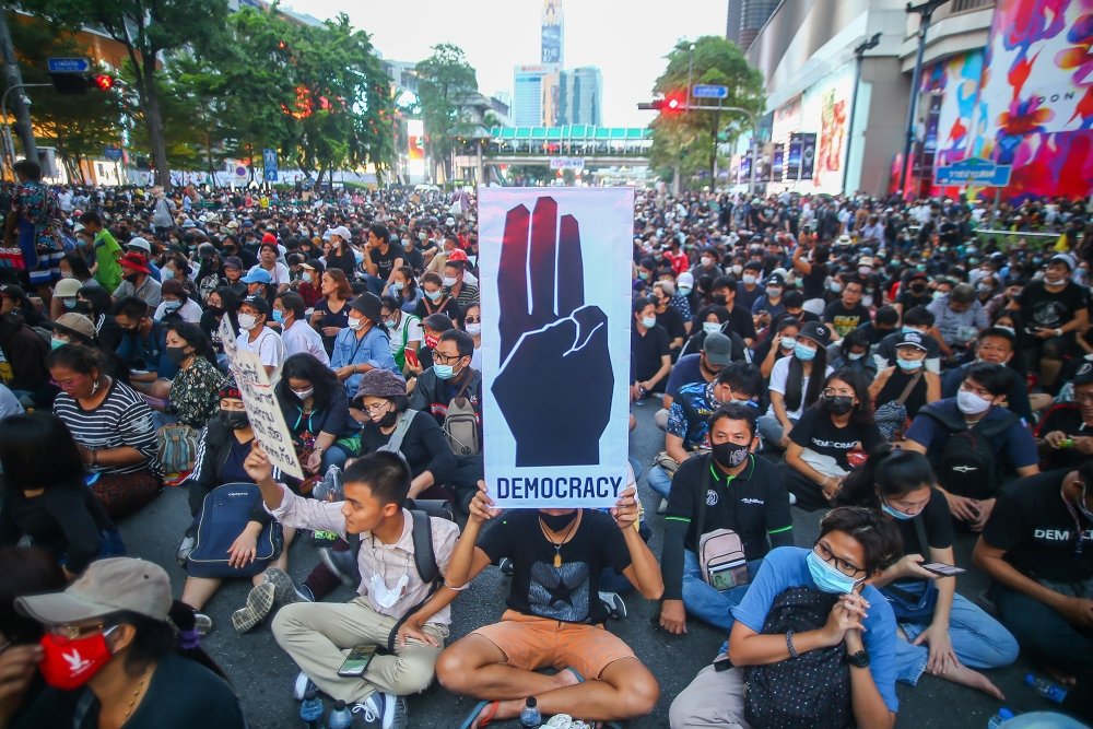 A crowd of demonstrators sit in a roadway, with one in the center holding a sign that reads "democracy"