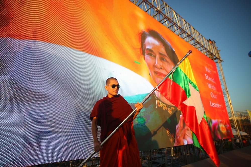 A man in red robes stands with a flag in front of a banner of Aung San Suu Kyi