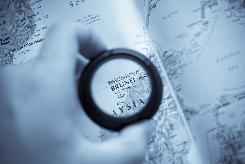 A hand holds a magnifying glass that is circling Brunei on a map.