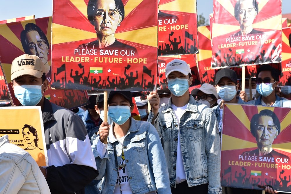 A crowd of protestors wearing surgical masks carry signs reading Please Save our Leader - Future - Hope with a photo of Aung San Suu Kyi