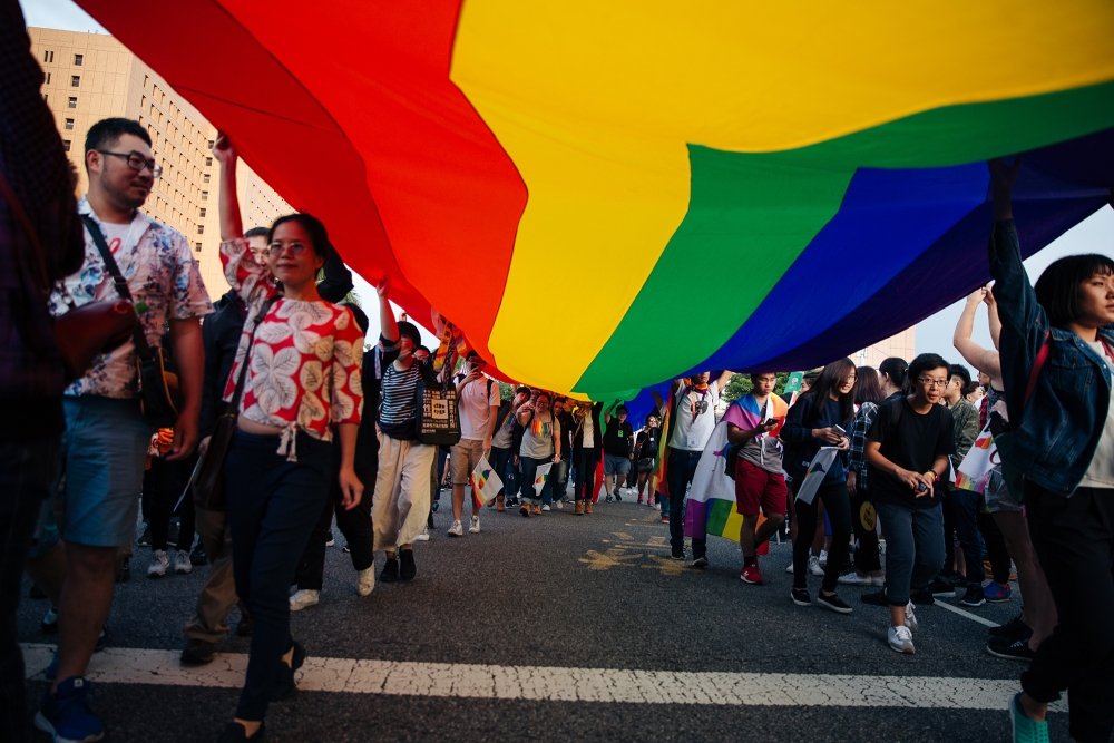 A group of people march carrying a large rainbow flag above their heads.