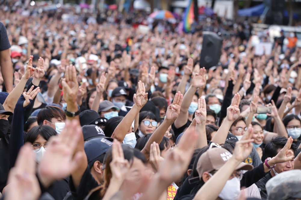 A crowd of people gathered at a protest, lifting their hands with three fingers extended in a gesture of protest against the government.
