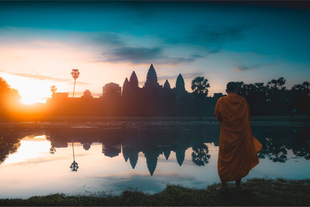 A monk stands in the foreground looking out at a temple at sunrise.