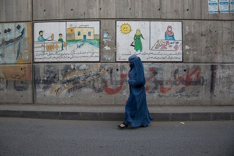 A woman in a blue burqa walks along an empty street in front of murals and graffiti.