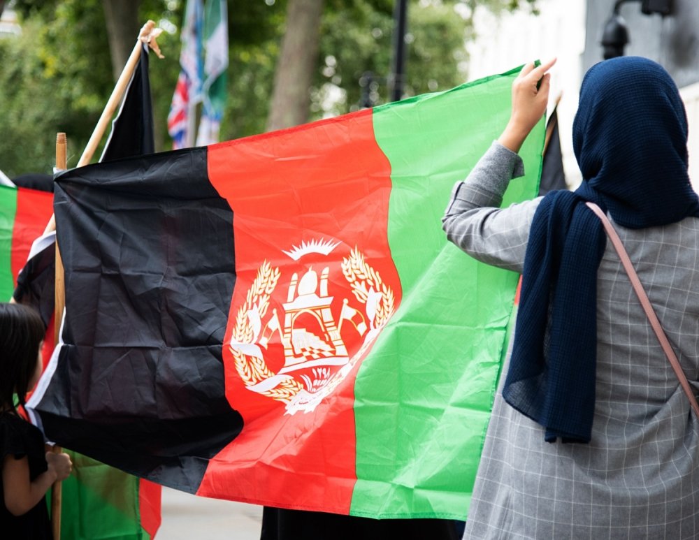 Two people with their backs to the camera are holding a large Afghan flag.