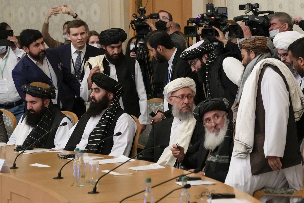 A group of Taliban officials sitting at a conference table with press behind them.