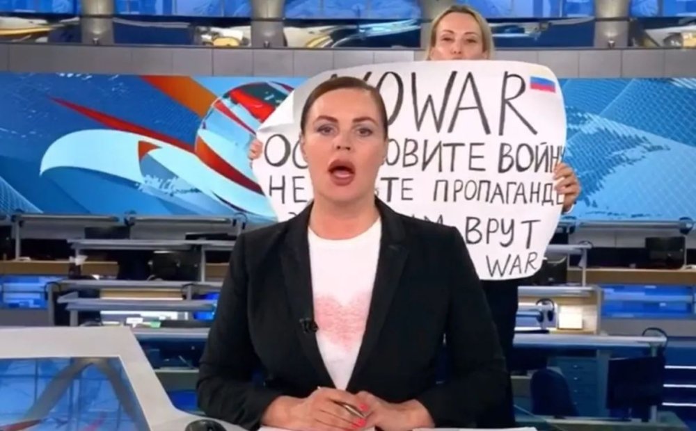 A protester holding sign behind a newscaster