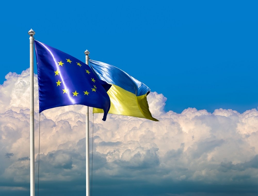 Two flags of Ukraine and the European Union on flagpoles at blue cloudy sky.