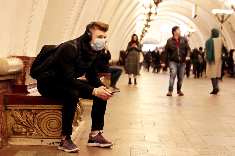 A young man with a backpack sits at a Moscow metro station with a medical mask