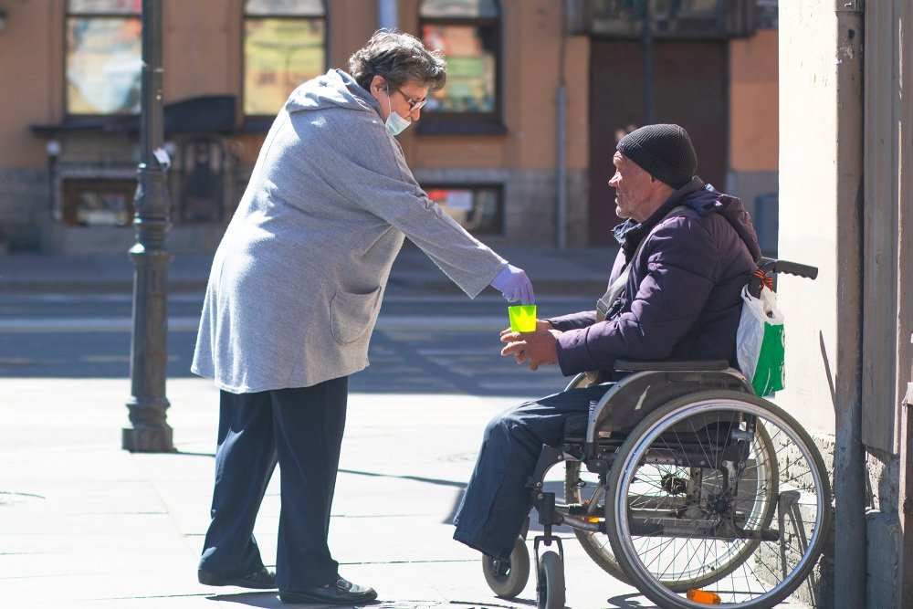Adult elderly woman in a protective medical mask and gloves gives money to a man in a wheelchair in St. Petersburg.