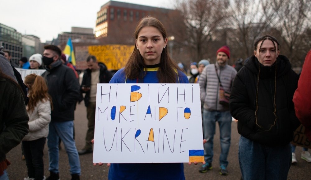 Young Person holding sign with text "Ні війні More Aid To Ukraine"