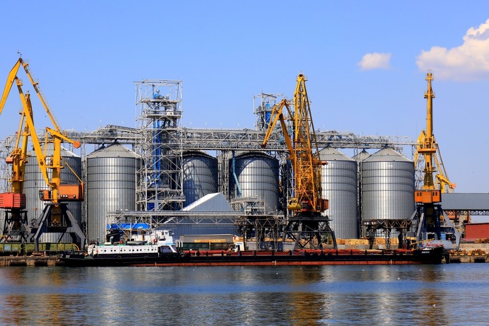 Harbor cranes unload barge at Odesa Seaport amid large metal tanks with wheat, grain, food. 