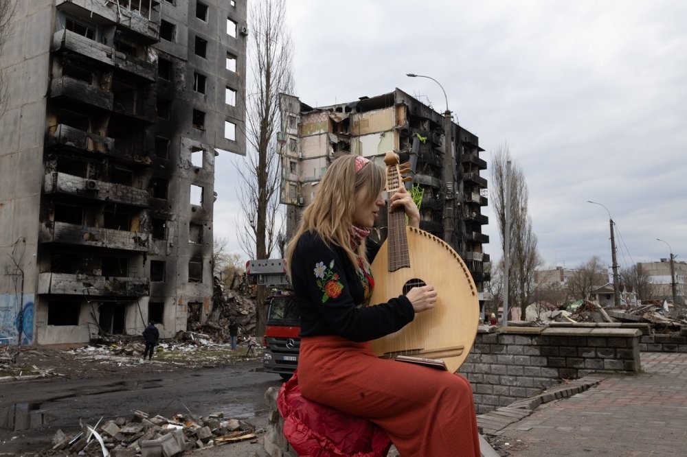 A woman plays a stringed instrument in front of a destroyed building in Bodoryanca, Kyiv Oblast, Ukraine