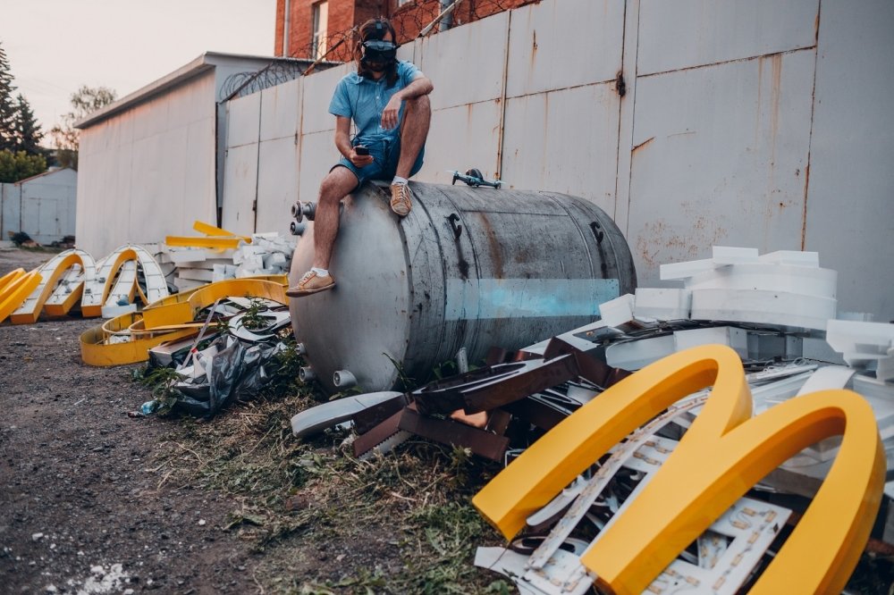  Young man sitting on VR headset and McDonalds logo "M" on dirty rubbish heap dump trash litter garbage.