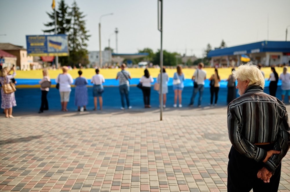 An older man observes a group of people holding a large Ukrainian Flag