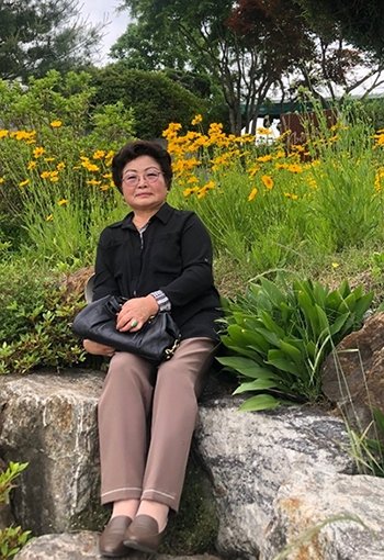 A Korean woman sits on a low wall in front of a field of yellow flowers.