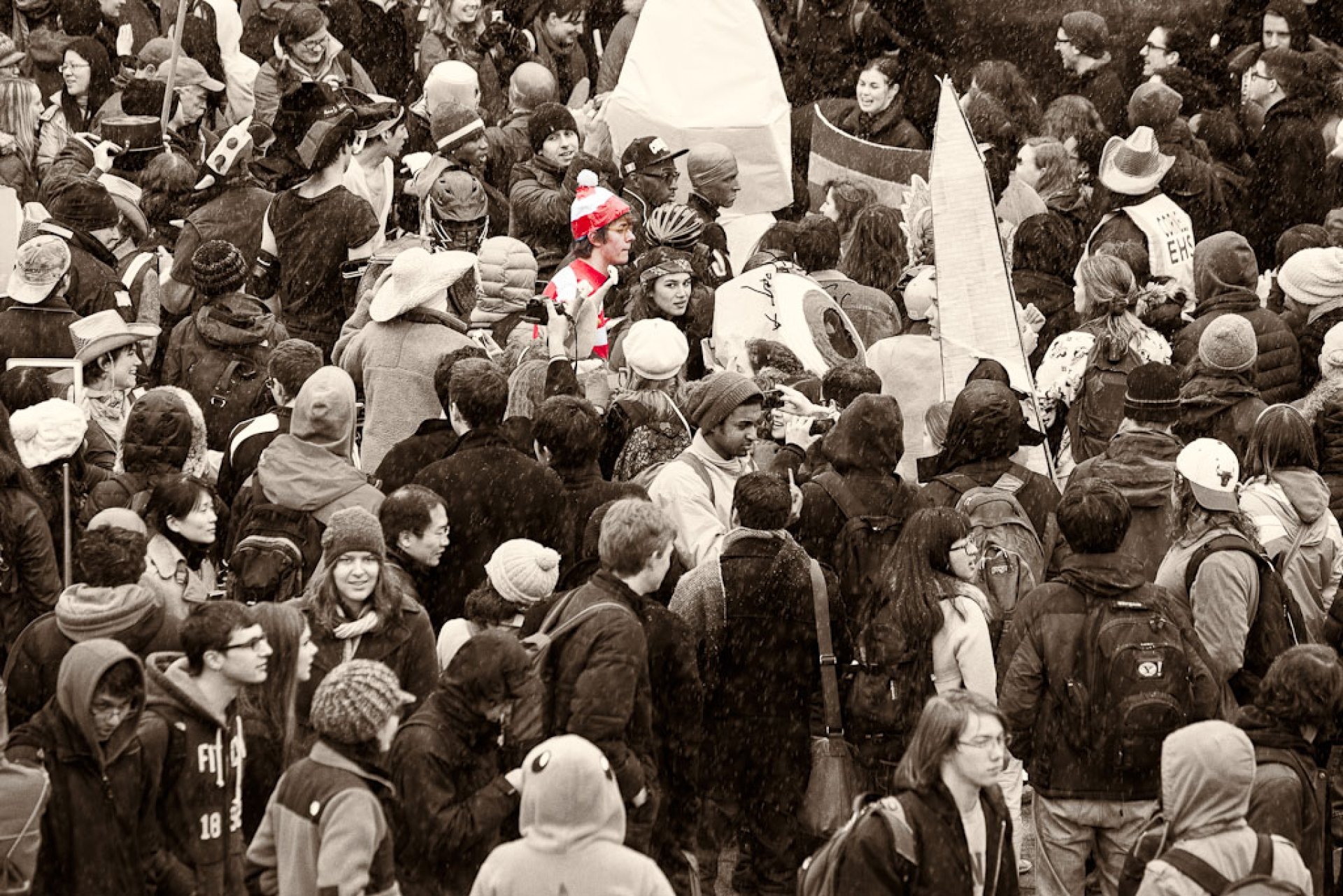 A photo in sepia of a crowd with a man wearing glasses and a red and white stripped sweater and had in color. 