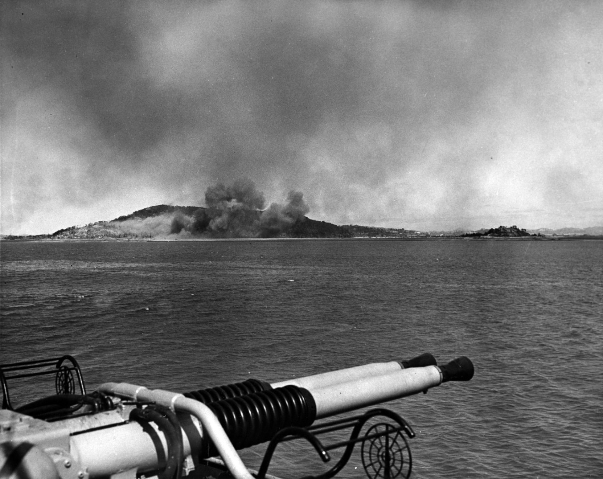 A black and white image of an island with smoke rising in the distance.