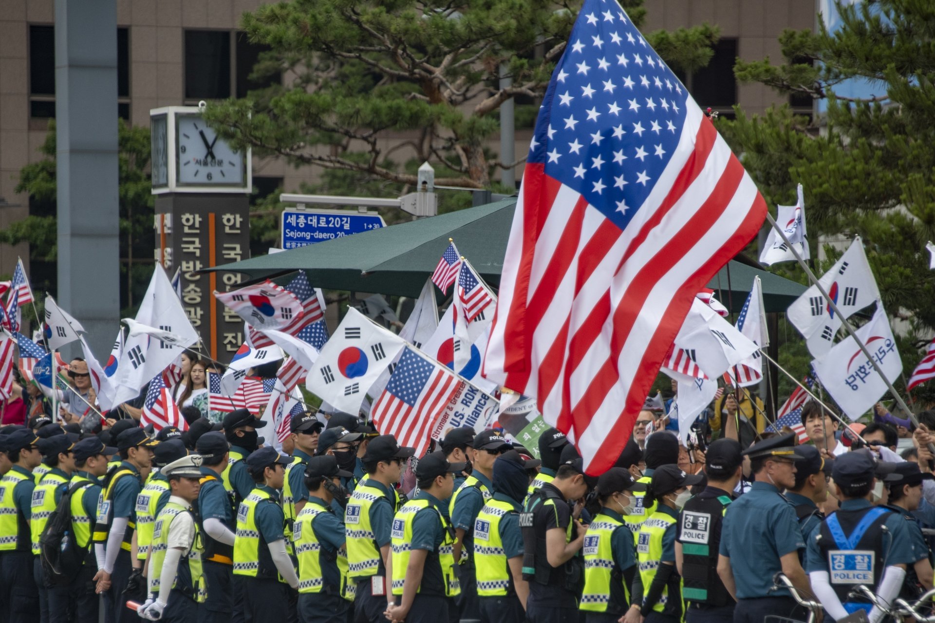 A crowd of people are waving U.S. and South Korean flags, standing behind a line of policemen.