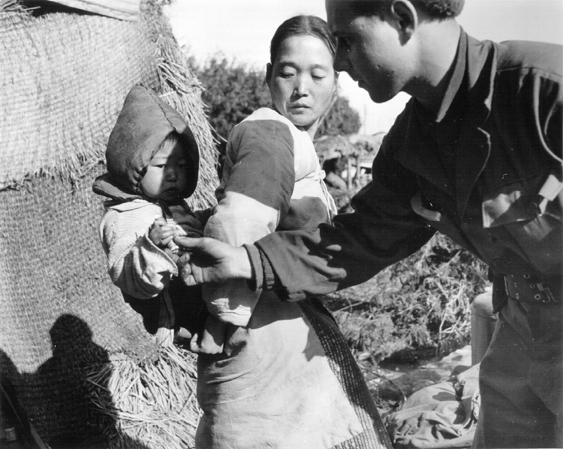 A black and white photo of a Korean woman standing with her child, and a U.S. soldier who is giving the child a piece of candy.