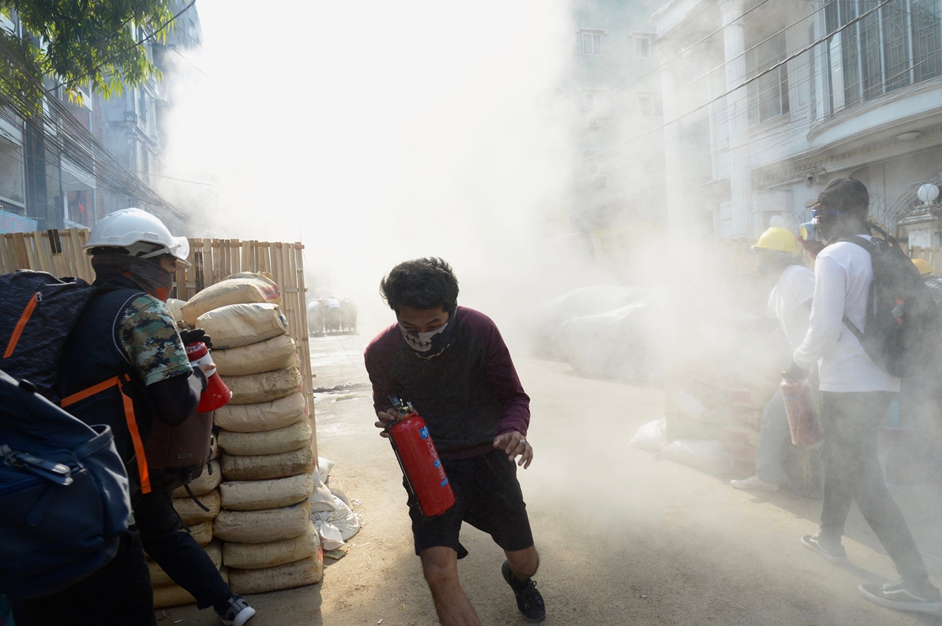 Several protestors in masks, carrying fire extinguishers, running from a cloud of smoke.