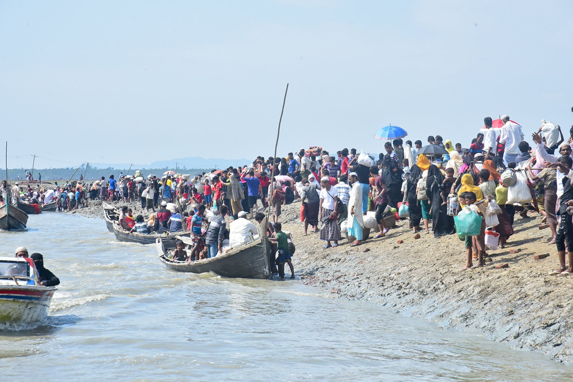 At a shoreline, there is a crowd of people on the beach and also a line of crowded boats with Rohingya refugees.