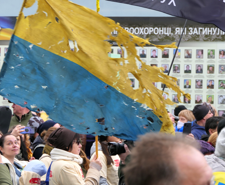 a tattered Ukrainian flag flies above a crowd of people