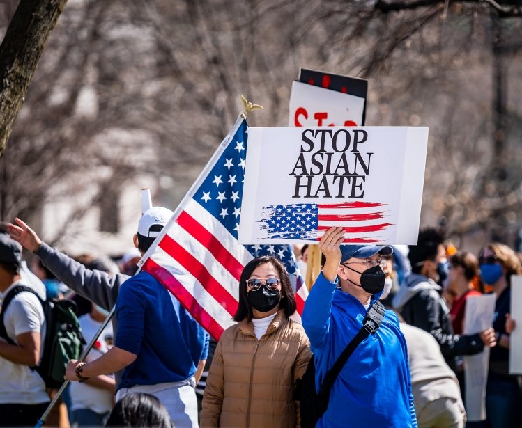 A man stands in a group of protestors wearing a masks, holding a sign with an American flag on it that says Stop Asian Hate.