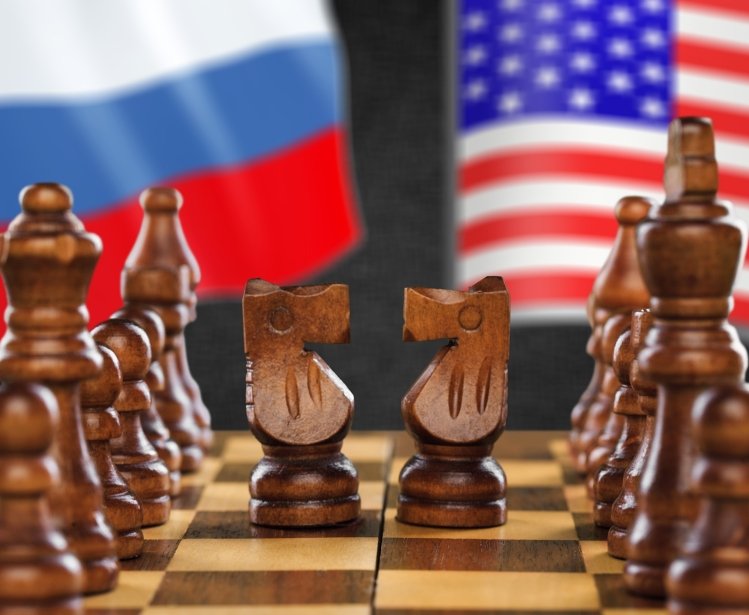 A chess board is set up with two knights facing each other, Russian and U.S. flags in the background.