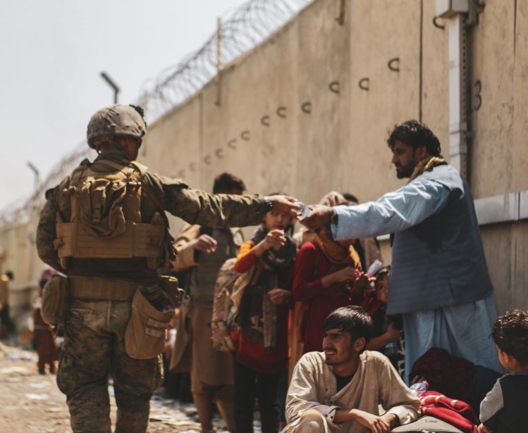A man in a military uniform hands a bottle of water to an Afghan civilian waiting in a crowd of people.