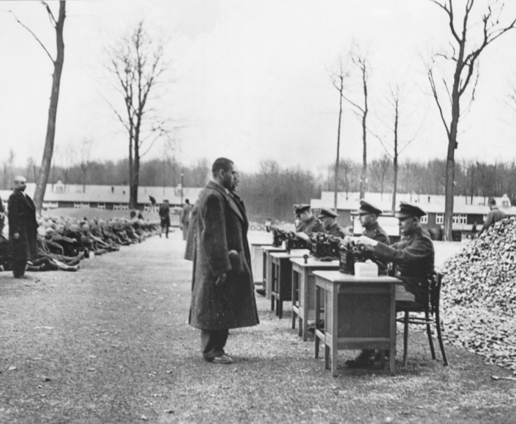 Gestapo officials recording data on incoming prisoners at a German concentration camp. Many others are seated on the ground