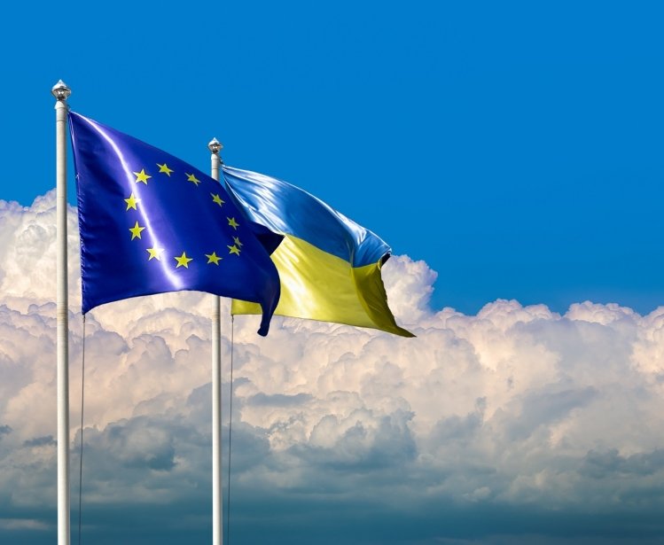Two flags of Ukraine and the European Union on flagpoles at blue cloudy sky.