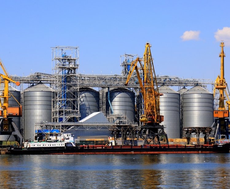 Harbor cranes unload barge at Odesa Seaport amid large metal tanks with wheat, grain, food. 