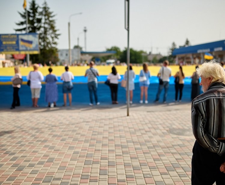 An older man observes a group of people holding a large Ukrainian Flag