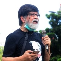 A photo of a bearded man speaking into a microphone at an ourdoor event.